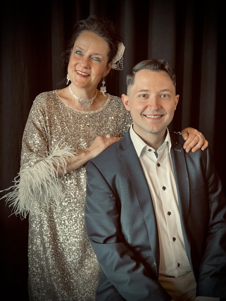 Ein Mann in Sakko sitzend, dahinter eine stehenden Frau im Abendkleid 