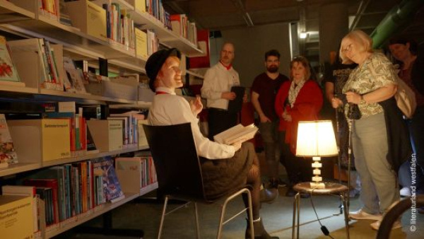 Szenenfoto: ein Mann sitzt vor einem Bücherregal, Publikum interagiert