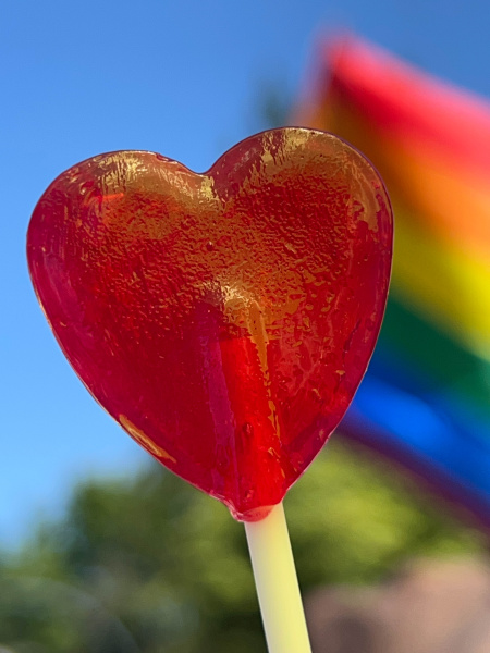Roter Herz-Lollipop im Vordergrund. Im Hintergrund sieht man eine Pride Flagge.